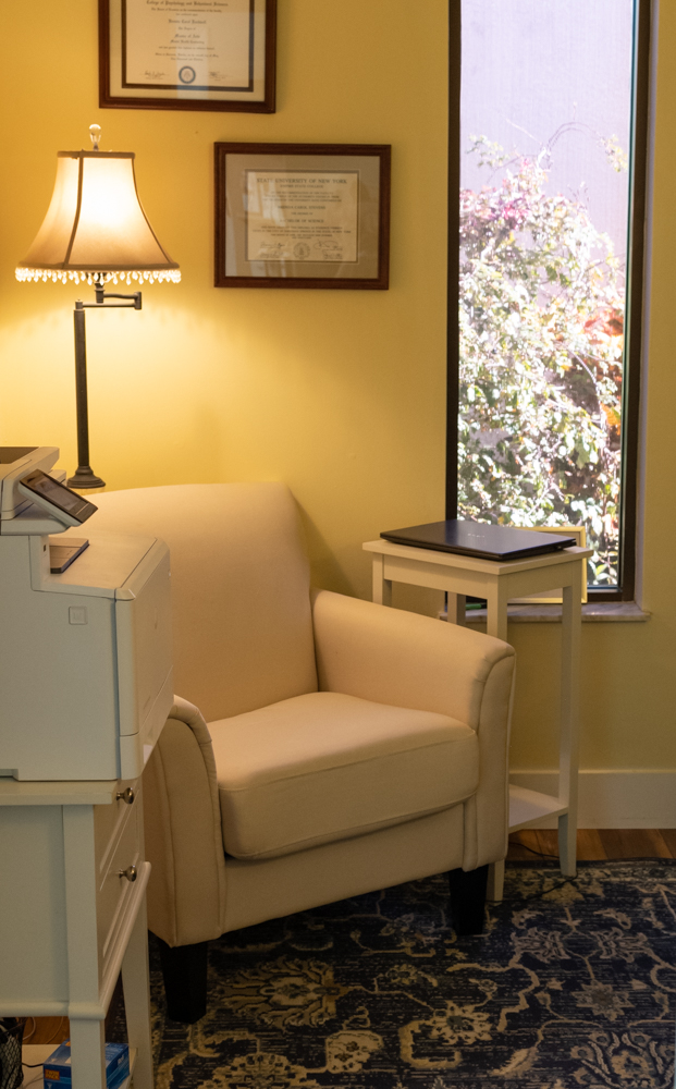 Cream colored chair next to printer and window with a lamp in the background and University degrees on the wall.